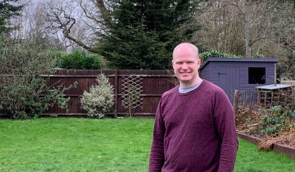 John Kilbey standing outside in a back yard with a back fence and shed.