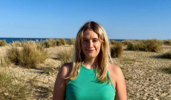 Josephine Bruford clos-up with a sandy beach with tall grasses in the background - ocean and blue sky in the distance.