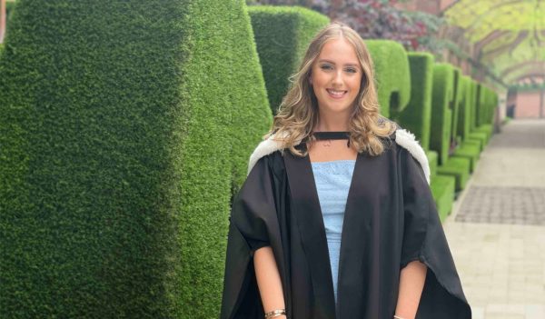 Megan Burn in a grad gown with manucured hedges in the background.