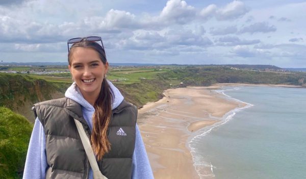 Mia McAfee with the view of a coastline behind her - green cliffs, sandy beach and the ocean.