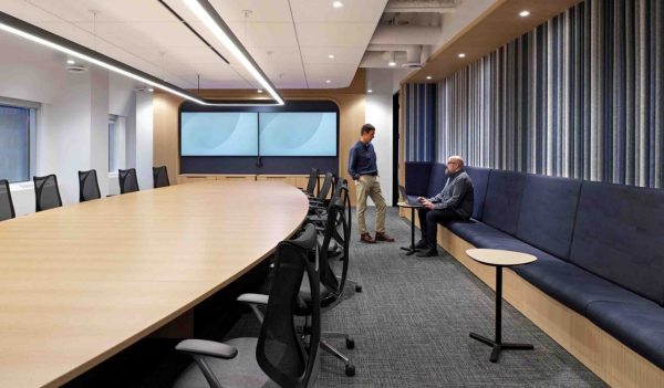 Interior conference room with large oval table, bench seating along the lenght of the room and a large screen at the end.